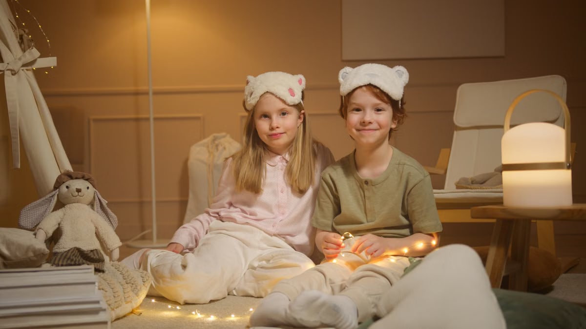 Two children in pajamas enjoy a cozy evening indoors with fairy lights and stuffed toys.